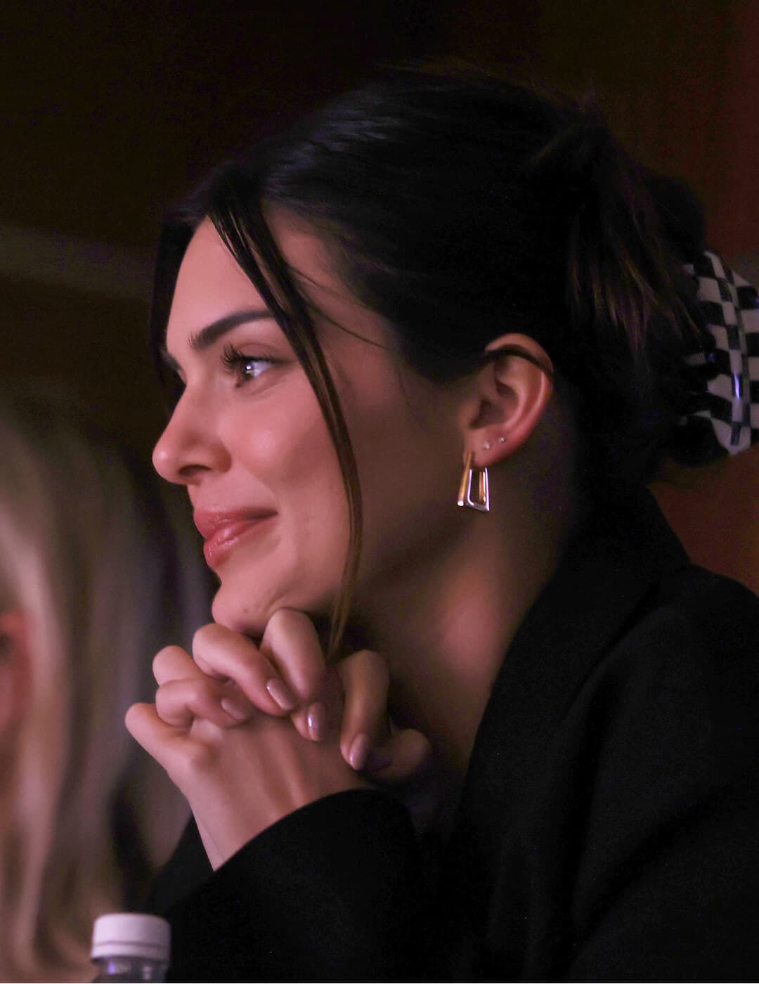 A photo of Kendall Jenner smiling with her hair styled in a claw clip updo wearing a black trench coat and gold earrings