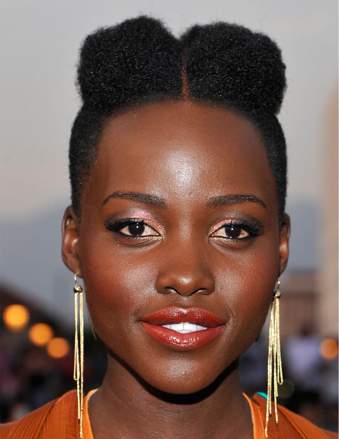 Close-up of Lupita Nyong'o wearing gold tassel earrings and orange dress rocking a faux mohawk hairstyle, shimmery coral eyeshadow and glossy coral lipstick makeup look