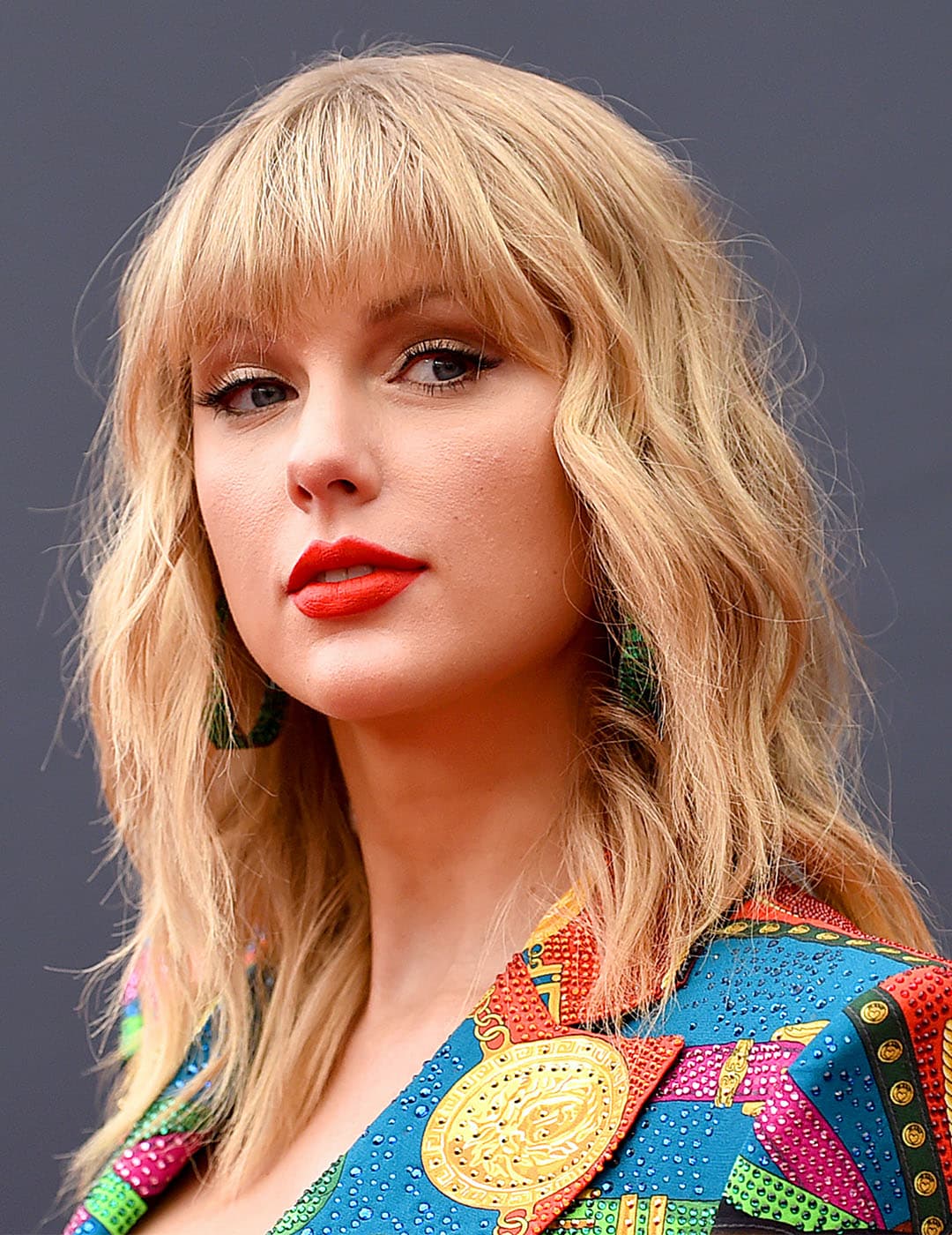 A photo of Taylor Swift flaunting her blonde shaggy hair paired with green hoop earrings and colorful top on a dark gray background