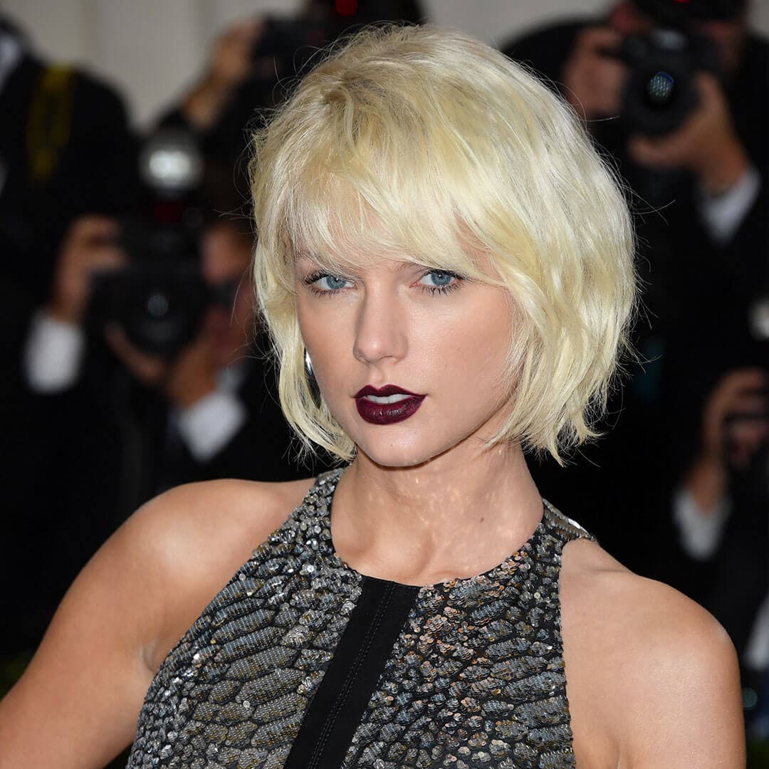 An image of Taylor Swift featuring dark purple-red lips and short blonde hair, dressed in a black sequined tank top dress