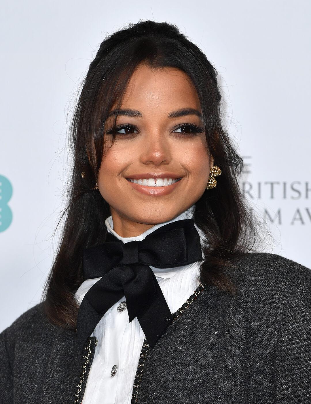 A photo of Ella Balinska wearing a bright smile in her nude lipstick, black bow tie, gold earrings with her black hair pulling a half-up, half-down hairstyle 