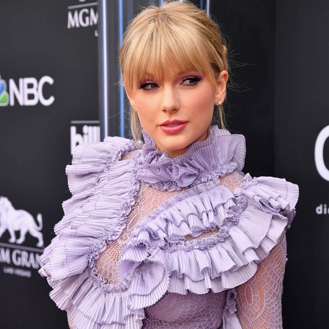 An image of Taylor Swift showcasing her Lavender Haze makeup look, dressed in a ruffled long-sleeve purple dress that features a slightly sheer mesh design