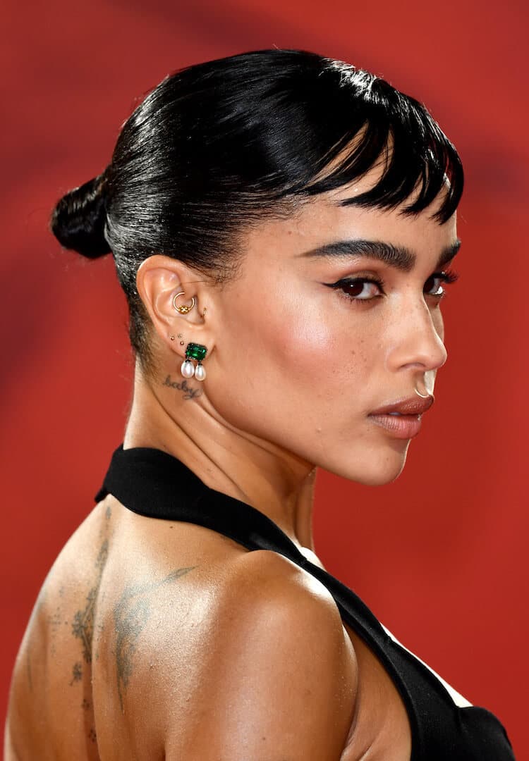 A close-up photo of Zoe Kravitz in a striking black dress revealing her bare back adorned with tattoos complementing her look with elegant pearl earrings and sports chic French-style baby bangs