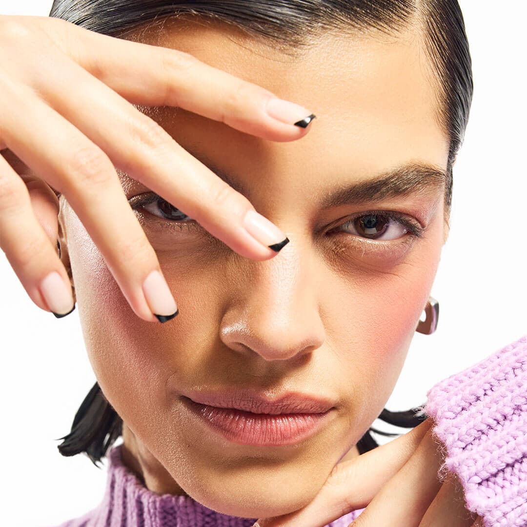 A close-up of a chic woman, her hand near her eye, highlighting her neatly shaped nude nails with sleek black tips