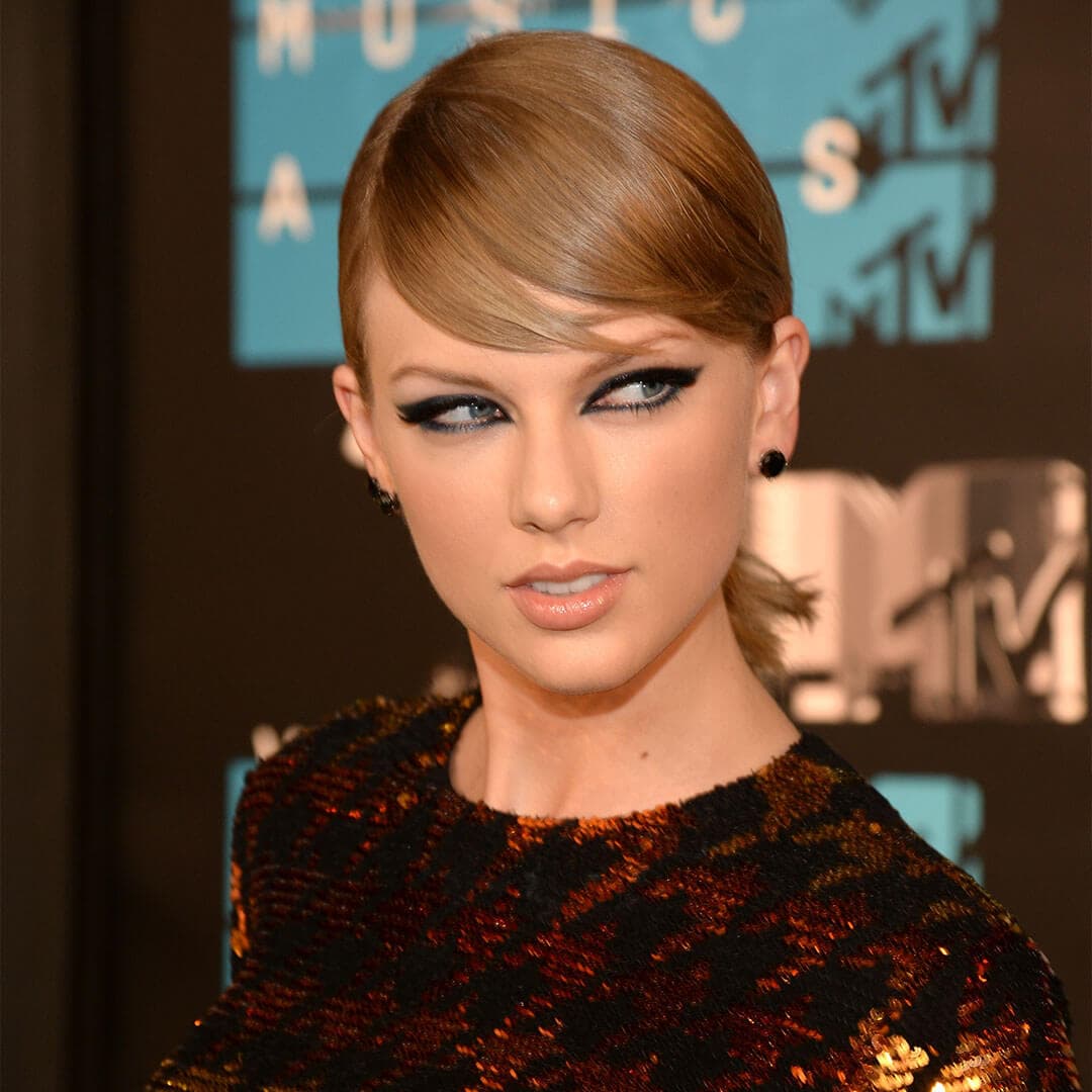 An image of Taylor Swift featuring bold eyeliner and nude lipstick, with her hair styled in slick, sideways bangs, wearing black earrings and a fitted long-sleeve top adorned with sequins