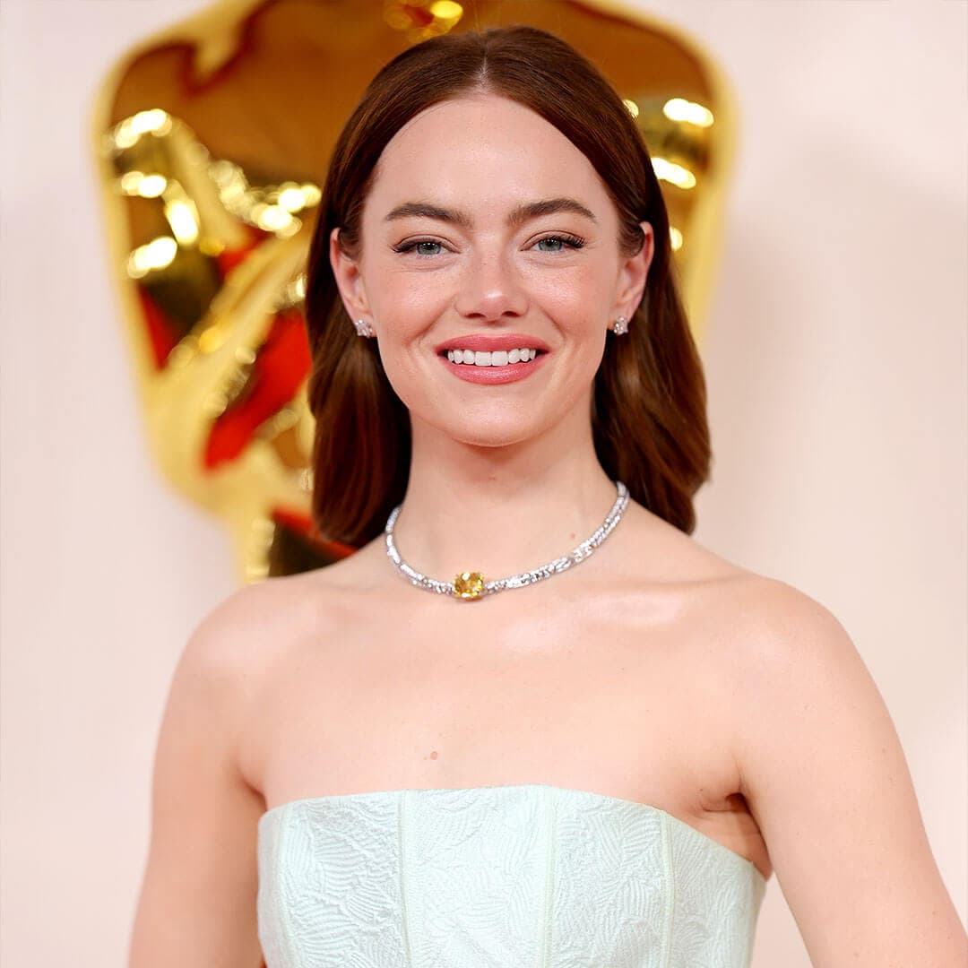 A photo of Emma Stone grinning at the camera while donning a pale pink monochromatic makeup and a white off-the-shoulder sheath gown