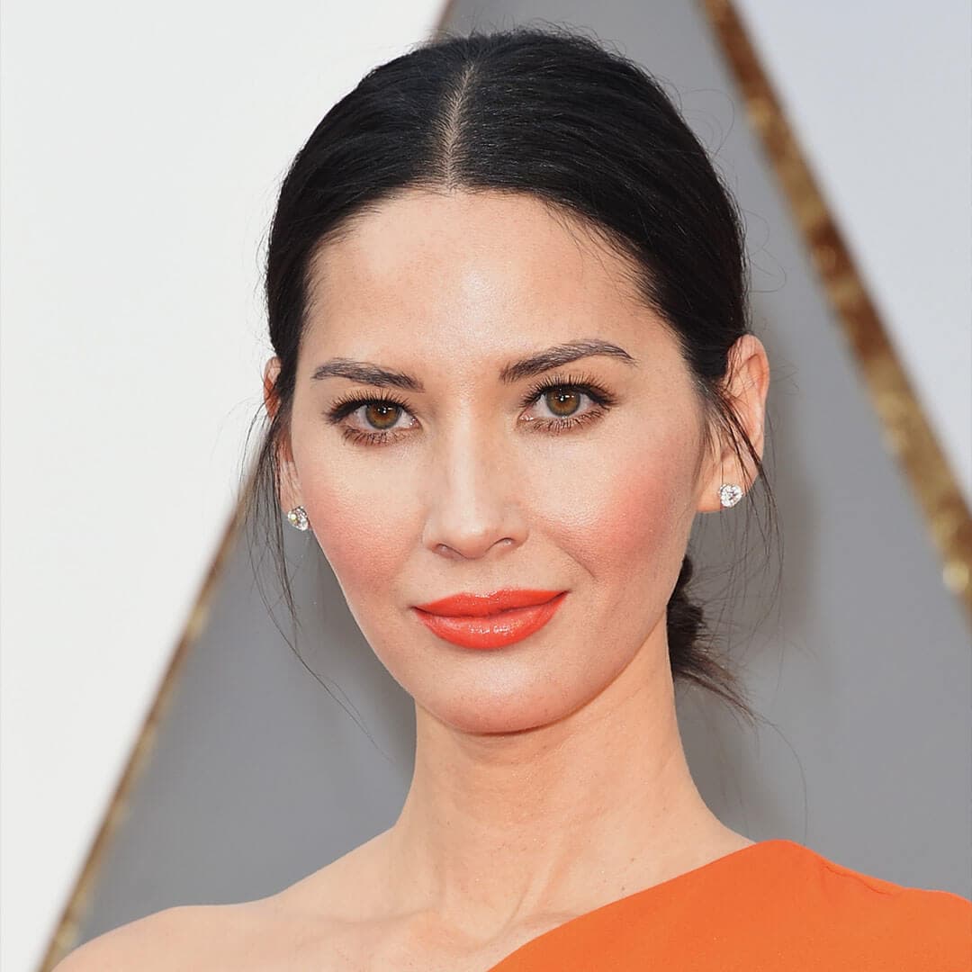 An image of Olivia Munn donning an orange off-the-shoulder dress, her black hair styled in a sleek low bun, and a fiery orange monochromatic makeup