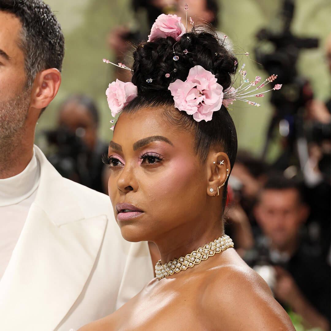 An image of Taraji P. Henson appeared in a minimal monochrome makeup look, with her hair done in an intricate top knot bun embellished with pink flowers, wearing a pearl beaded corset dress