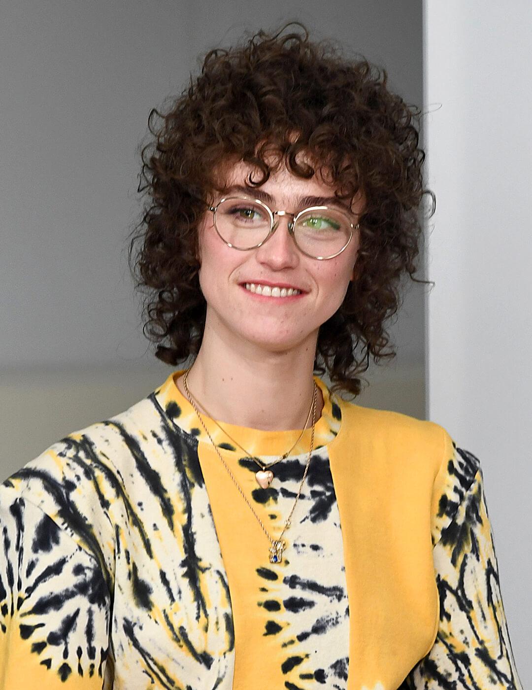 A photo of Ella Emhoff in her black curly mullet wearing a round-wired eyeglasses, yellow tie-dye T-shirt, and black pants