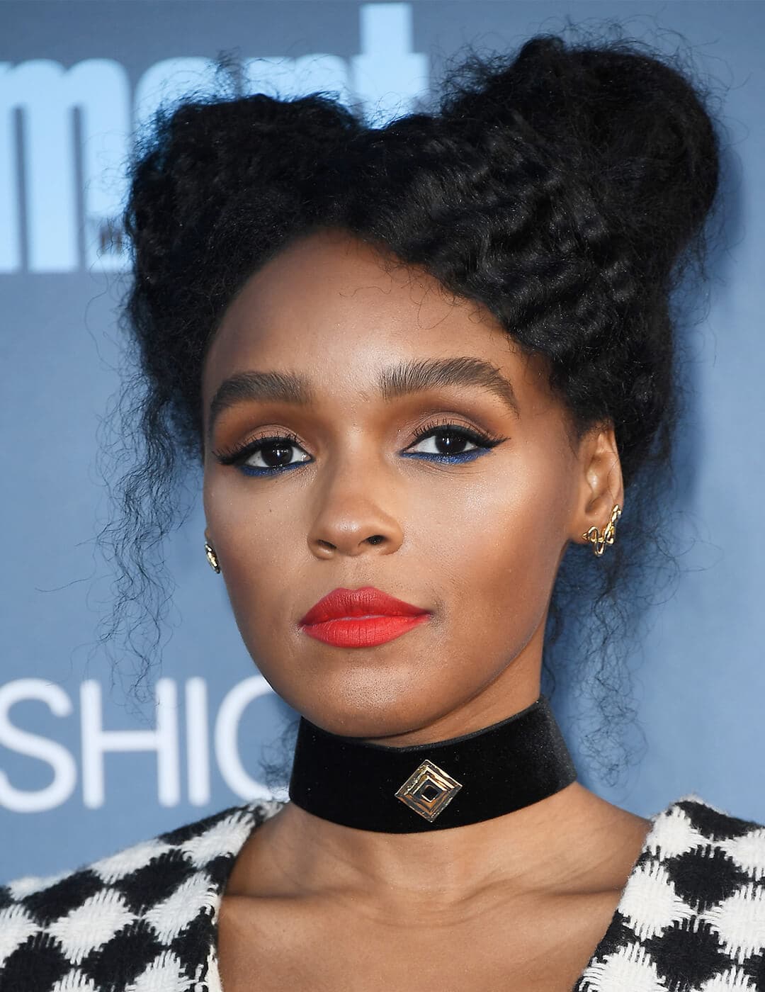 A photo of Janelle Monae styled her black curly hair into space buns wearing a black choker, a shade of red lipstick and a blue eyeliner