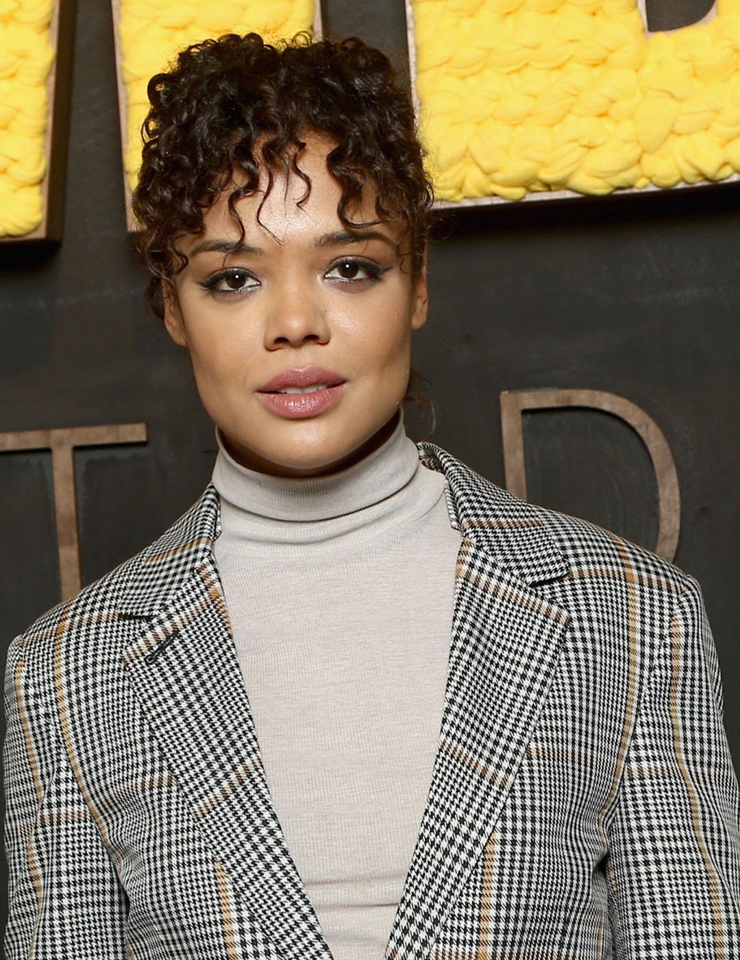 A photo of Tessa Thompson in her ivory-colored inner turtleneck topped with a stripe coat that accentuates her dark curly fringe hair on top of her face