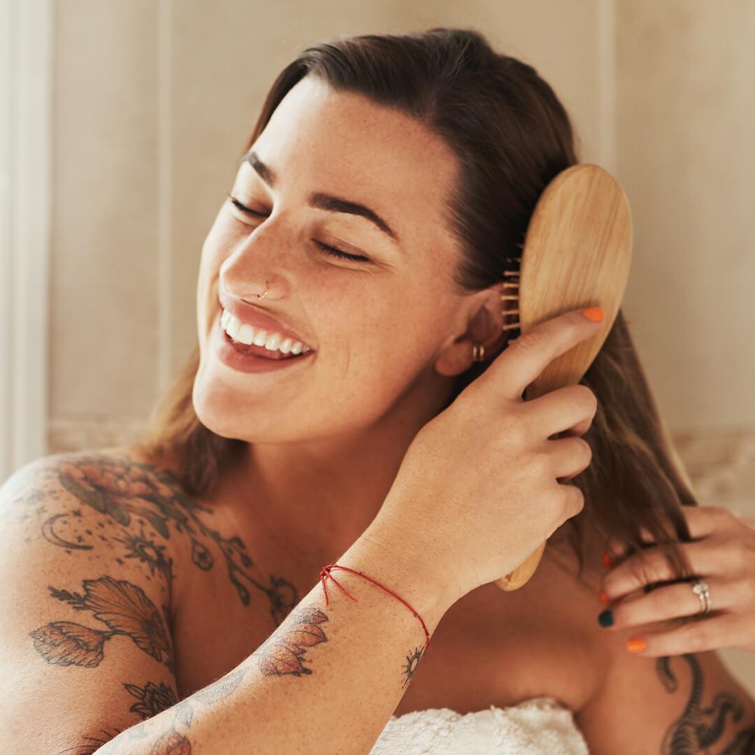 Woman with tattoos smiling and brushing her hair with a wooden brush