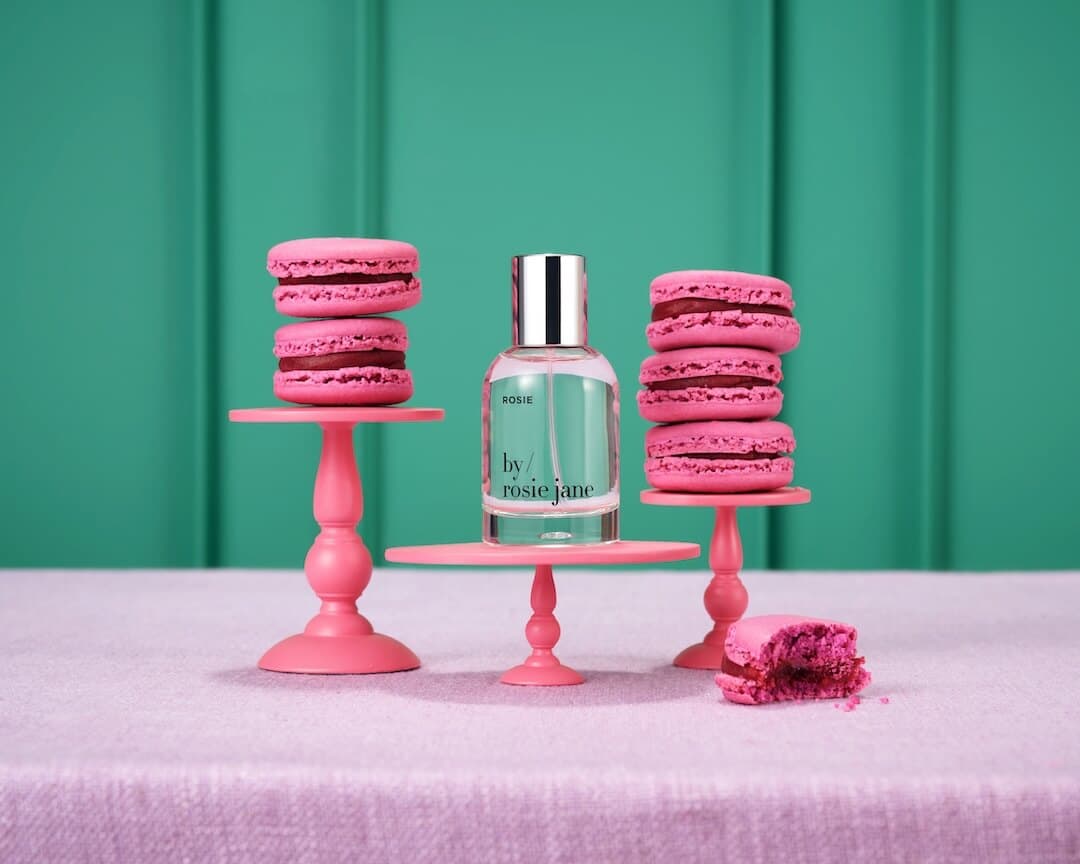 BY ROSIE JANE Rosie Eau de Parfum on a tiny pink cake stand next to two stacks of pink macaron desserts.