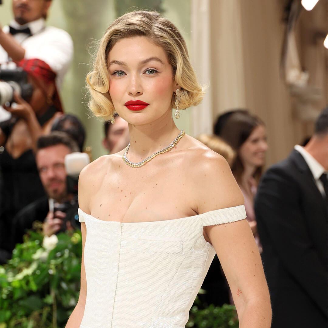 A photo of Gigi Hadid showing off her short blond hair, bright red lipstick, white off-the-shoulder dress, and diamond gold necklace with a monochromatic look