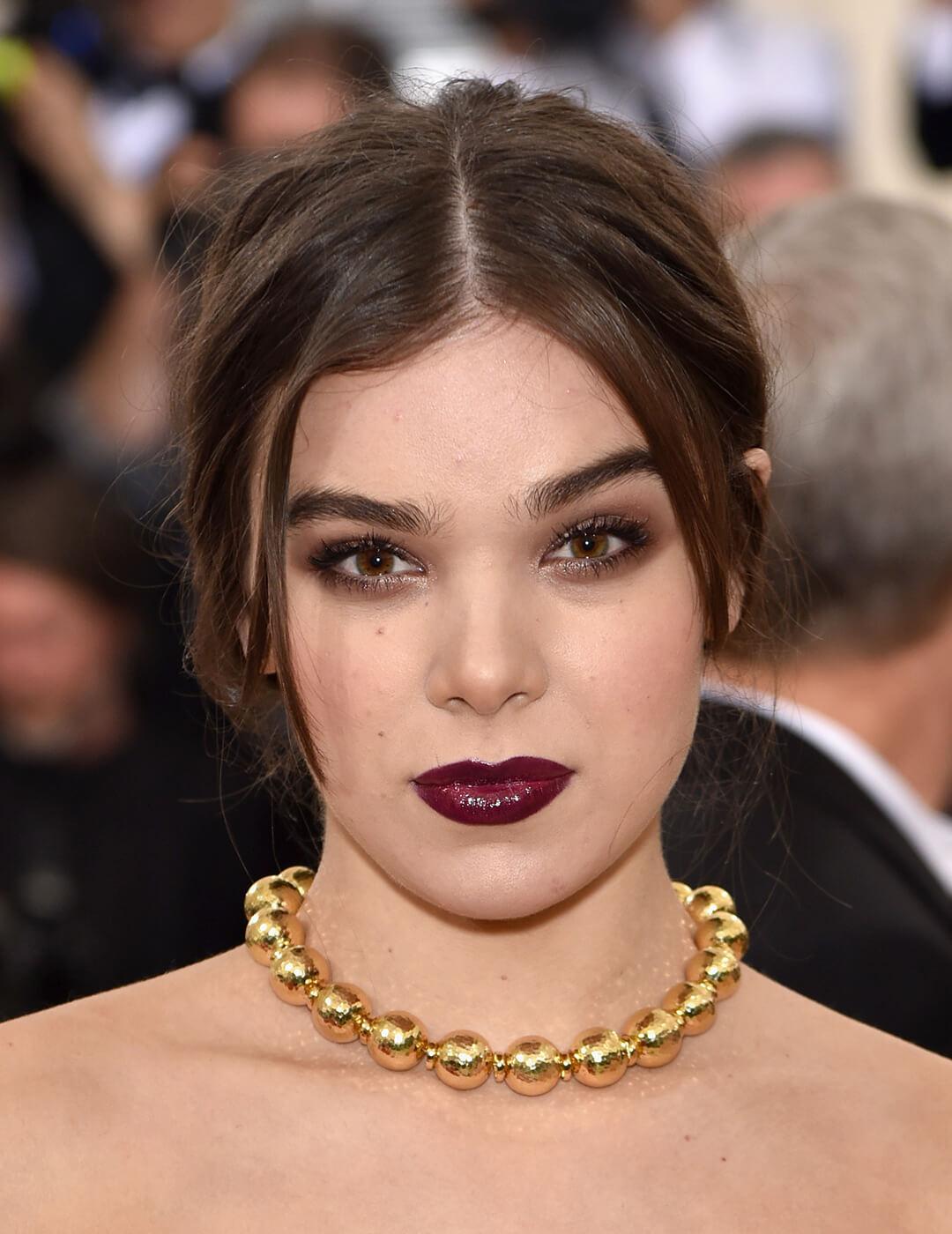 Close-up of Hailee Steinfeld rocking a smoky eye makeup look, glossy burgundy lips, and golden big ball chain necklace