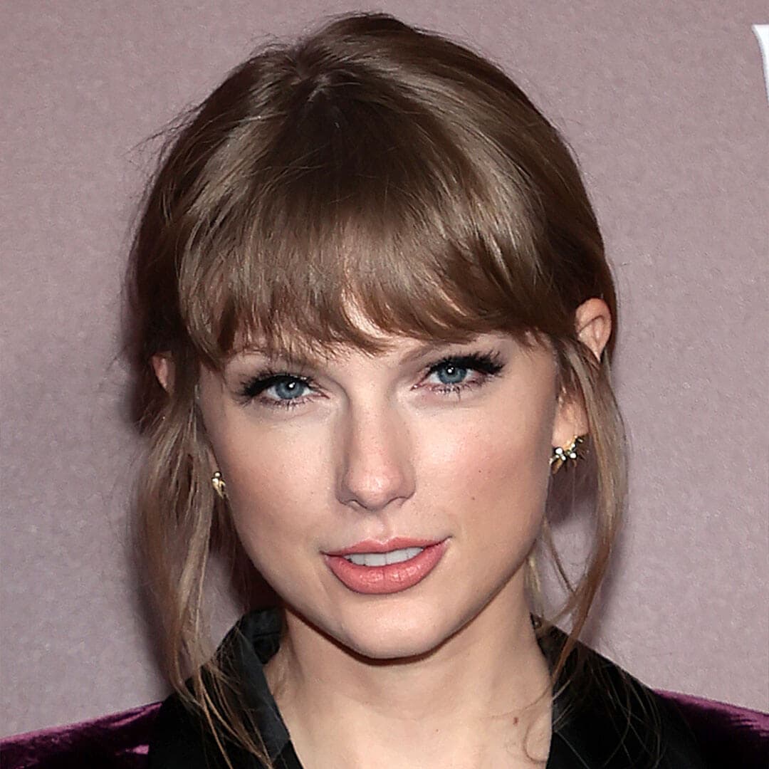 An image of Taylor Swift featuring her Feminine Fluttery Lashes makeup look, gazing directly at the camera while dressed in a velvet suit, her hair is styled with bangs and tied back