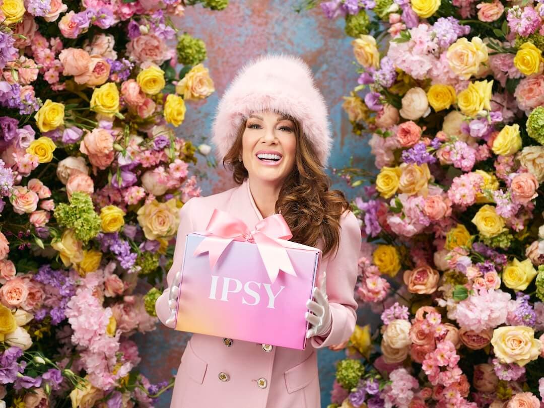 Lisa vanderpump in a pink suit and fluffy pink hat holding an Icon Box box with a bow on it and standing in front of a flower wall.