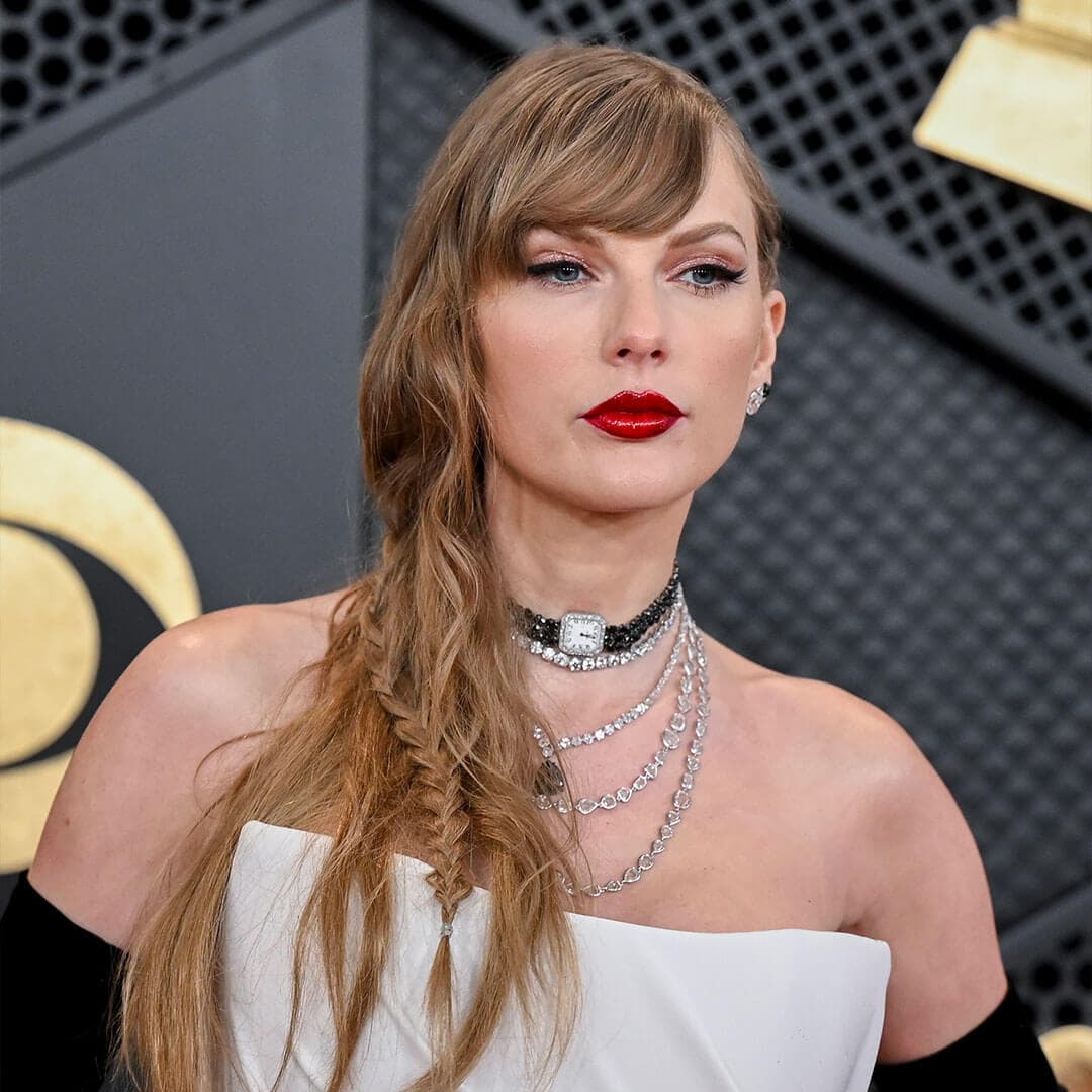 An image of Taylor Swift featuring her signature cat eyeliner and bold red lips, adorned with multiple gem necklaces, her hair is stylishly tucked down to the side, and she is wearing a white strapless dress along with black elbow-length gloves