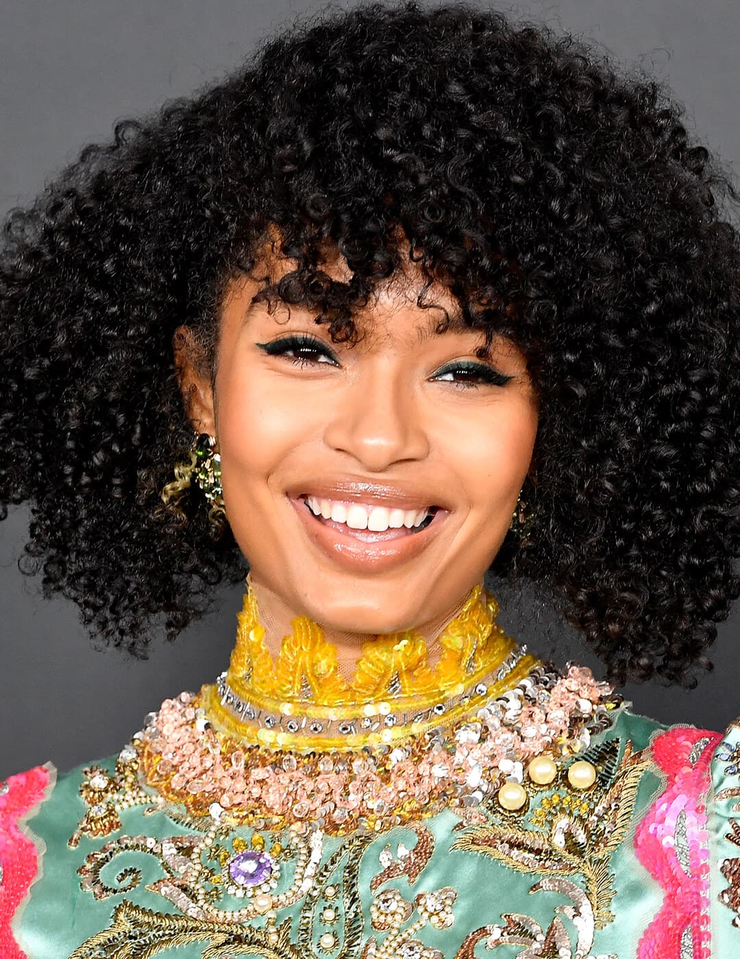 A photo of Yara Shahidi wearing a bright smile in her natural black curly hair with a curl specific haircut dressed in green full of pearls and beads 