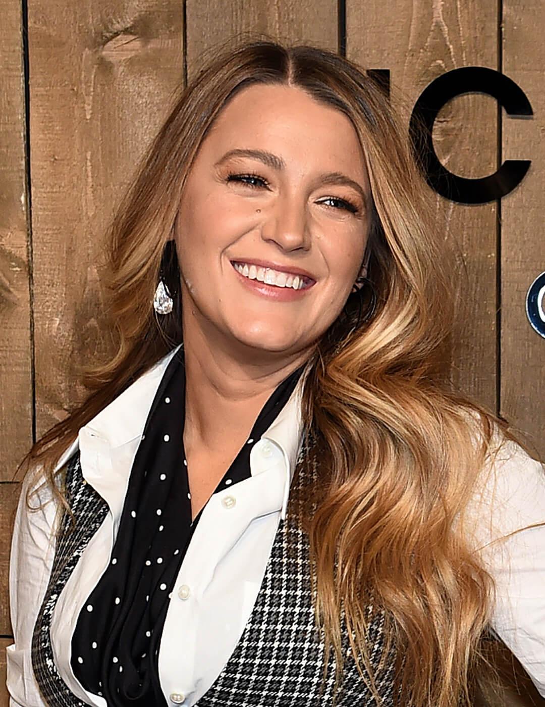A photo of Blake Lively in her blond loose waves wearing a neutral makeup , a black and white checkered vest with an inner white long sleeves 