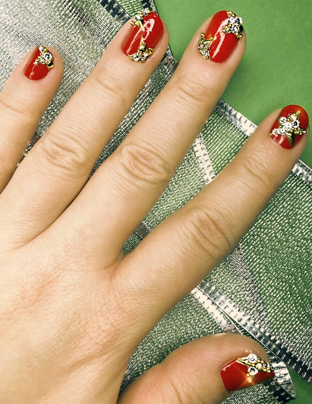 Close-up image of a model's hand with red mani embellished with rhinestones and studs against a green background with silver lace ribbons