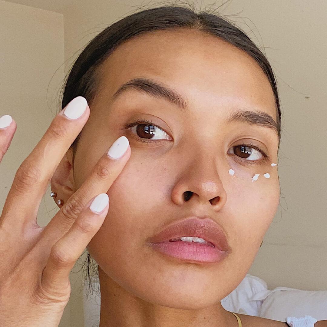 Close-up image of model with white mani putting on eye cream on clean skin