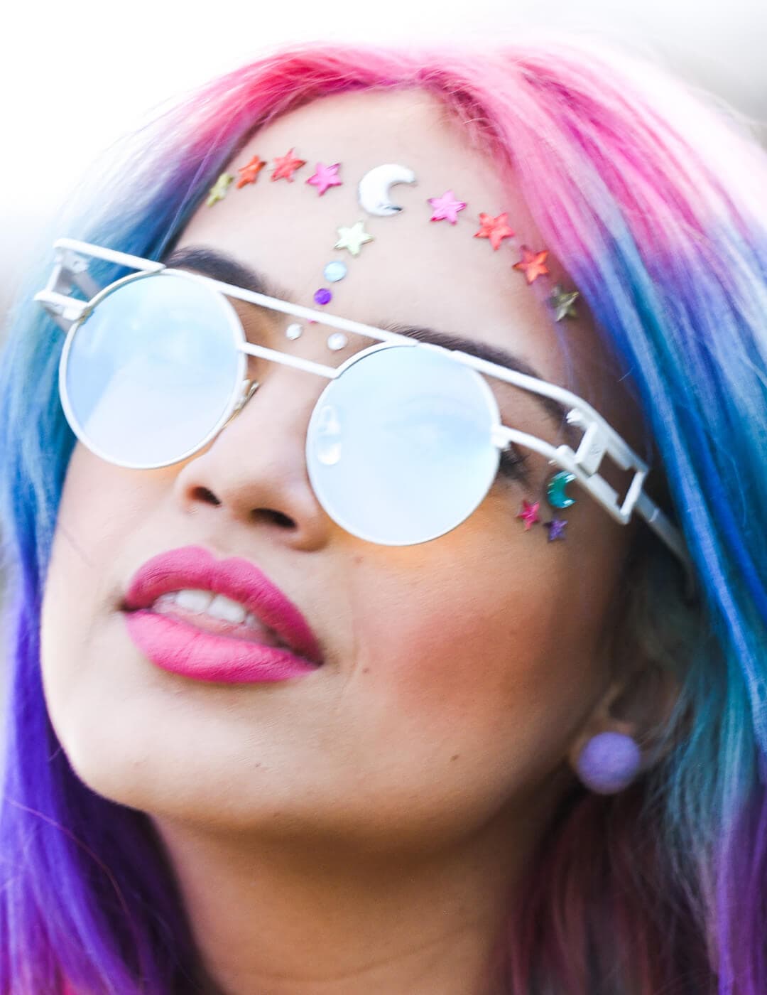 Close-up of a woman with rainbow hair color rocking sunglasses, hot pink lips, and face full of stars and moon gemstones