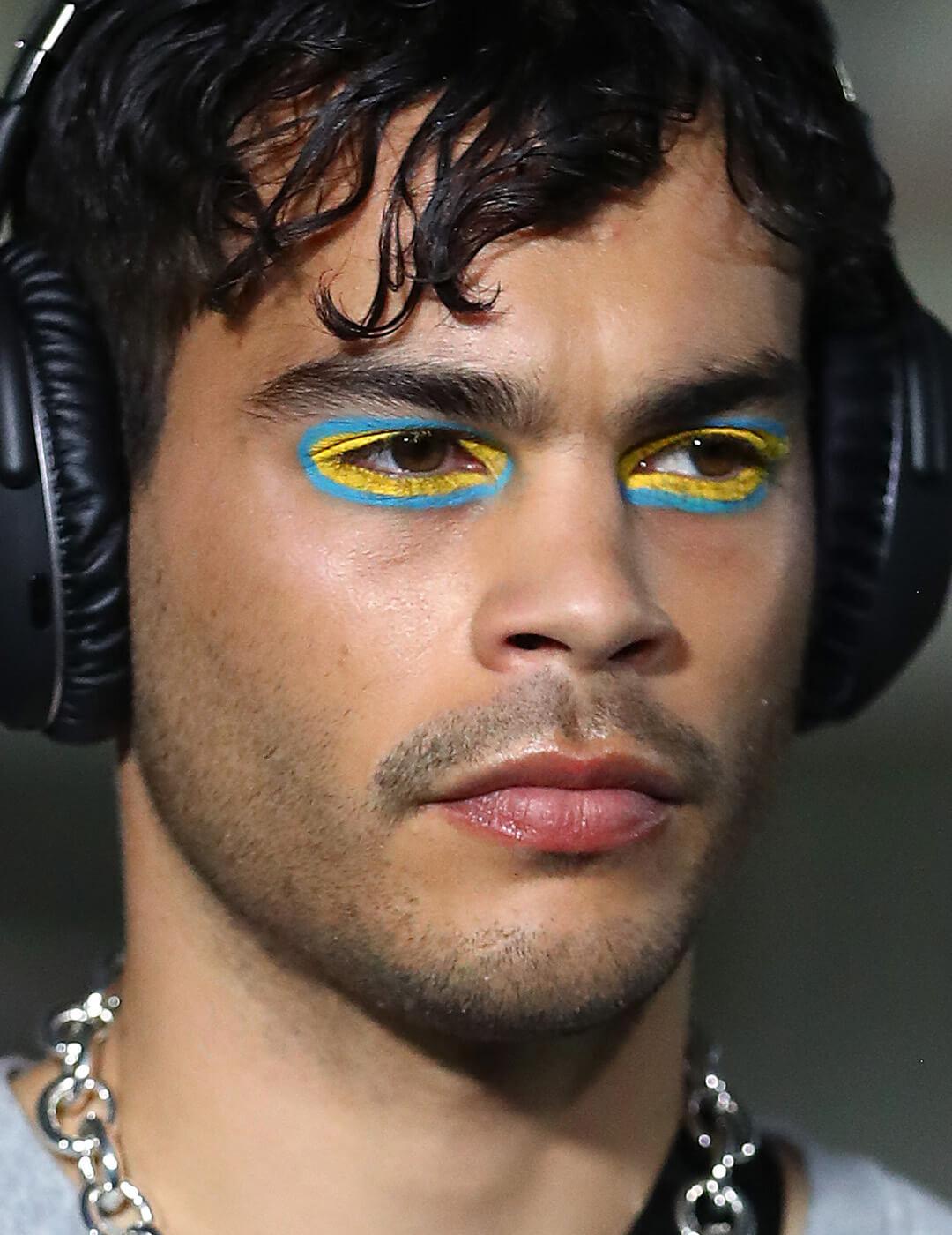 Male model wearing black headphones, silver necklace, and blue and yellow eyeshadow makeup