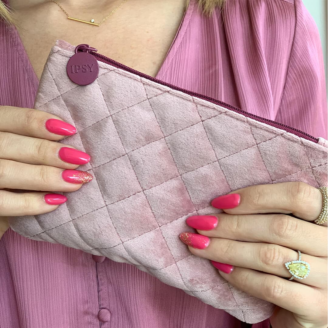 Close-up image of model's hands with fuchsia and gold glitter nail art holding IPSY February Glam Bag
