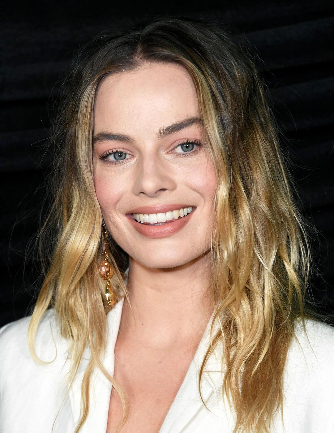 Margot Robbie wearing a white suit and loose waves hairstyle smiling