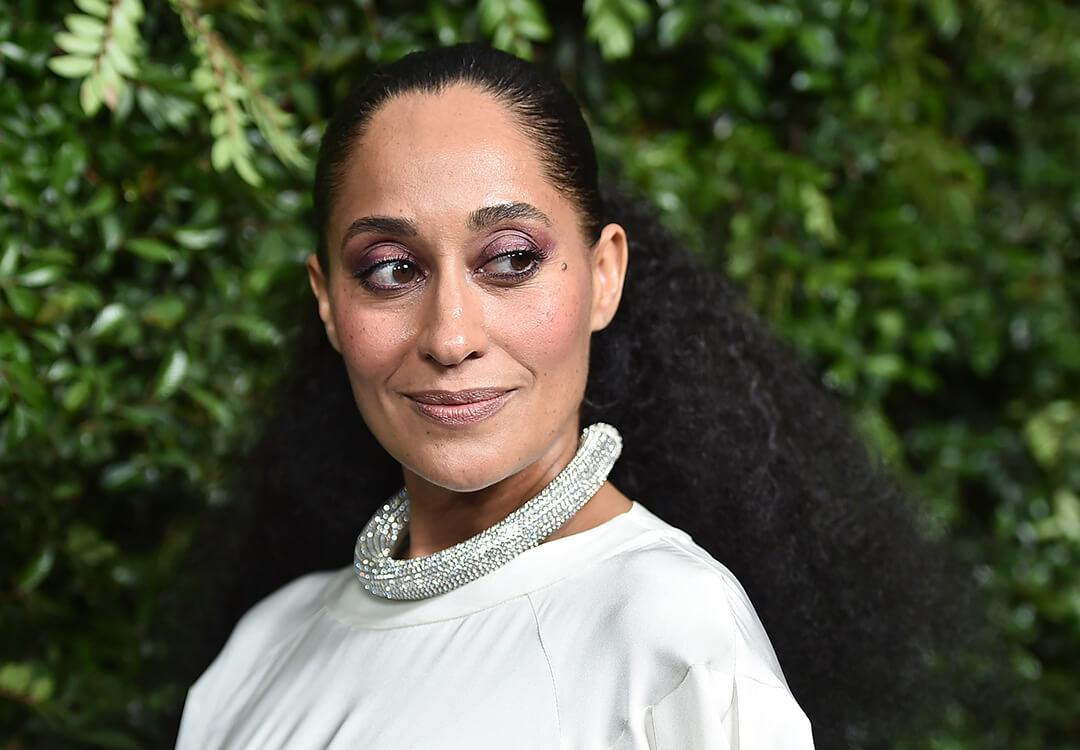 Tracee Ellis Ross in a silver dress rocking a dark purple eye makeup look, thick silver necklace, and a slicked back hairstyle