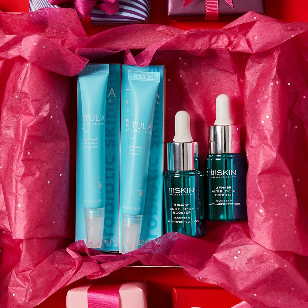 Flat lay image of two tubes of TULA SKINCARE Go Away Acne Spot Treatment and two bottles of 111SKIN 3 Phase Anti Blemish Booster inside a pink gift box on red background
