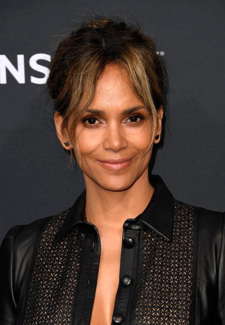 A close-up photo of Halle Berry wearing an open-buttoned black leather one-piece suit boasting curtain bangs hairstyle donned a chic black earring