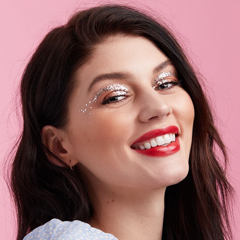 A close-up image of a model wearing nude eyeshadow with silver glitter gel arches paired with a bold red lipstick