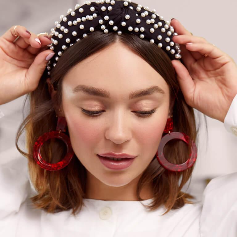 An image of a model with a no-makeup makeup look wearing bold red earrings while touching her pearl-studded headband