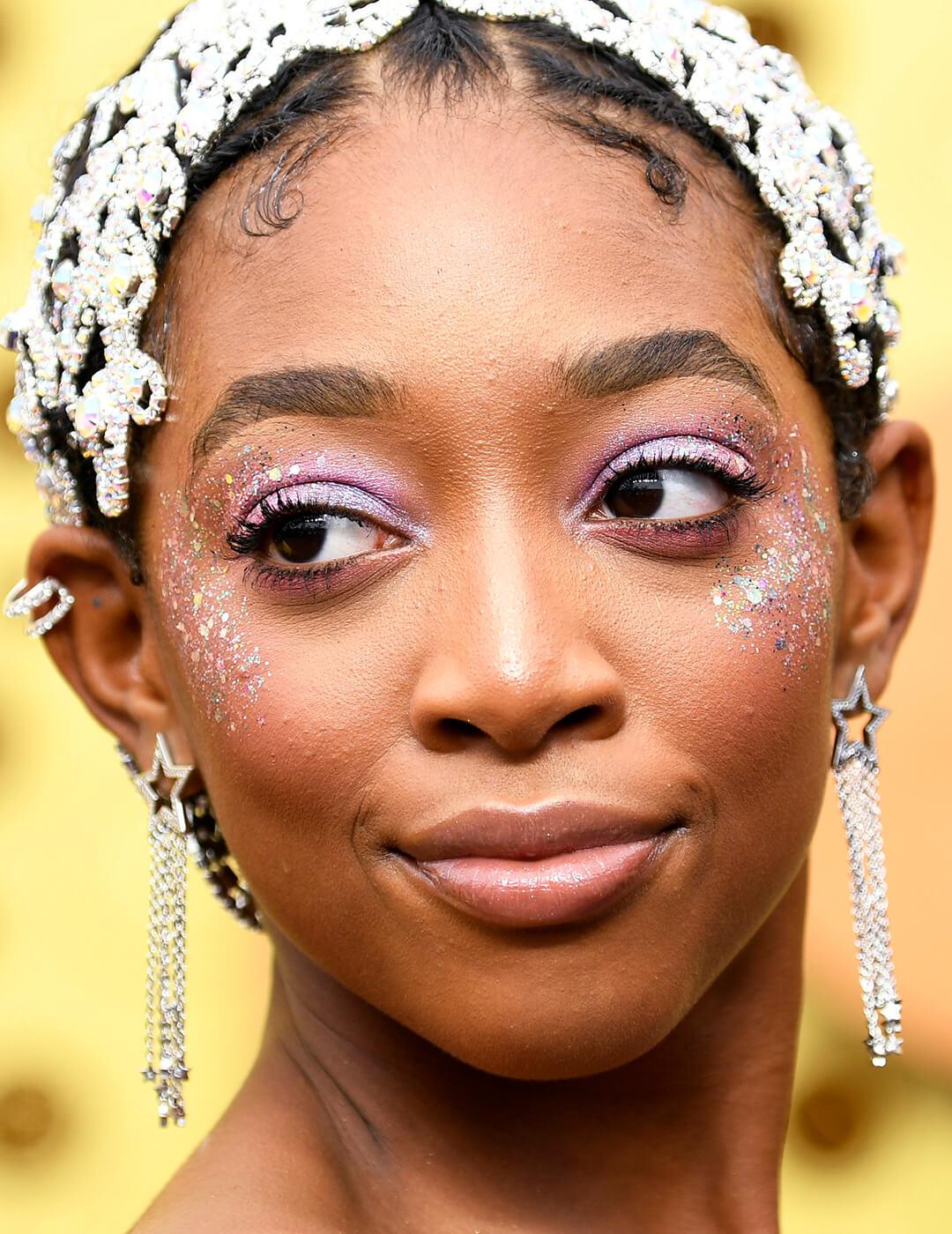 Close-up of Eris Baker rocking a shimmery pink eyeshadow look with colorful glitters on her cheeks, intricate silver headdress, and silver dangling earrings