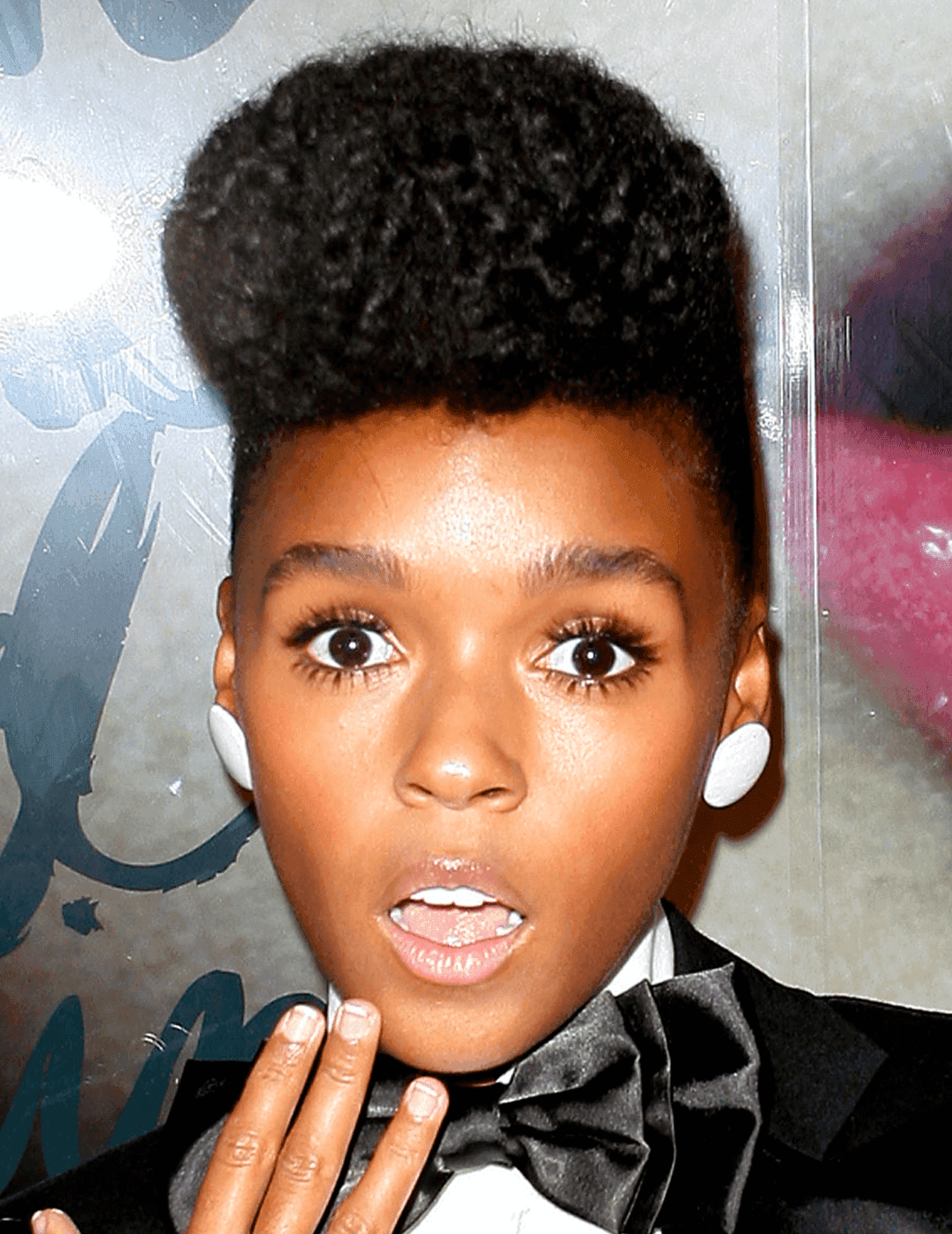 Janelle Monae in a quiff hairstyle, black suit and blow, and white stud earrings looking shocked
