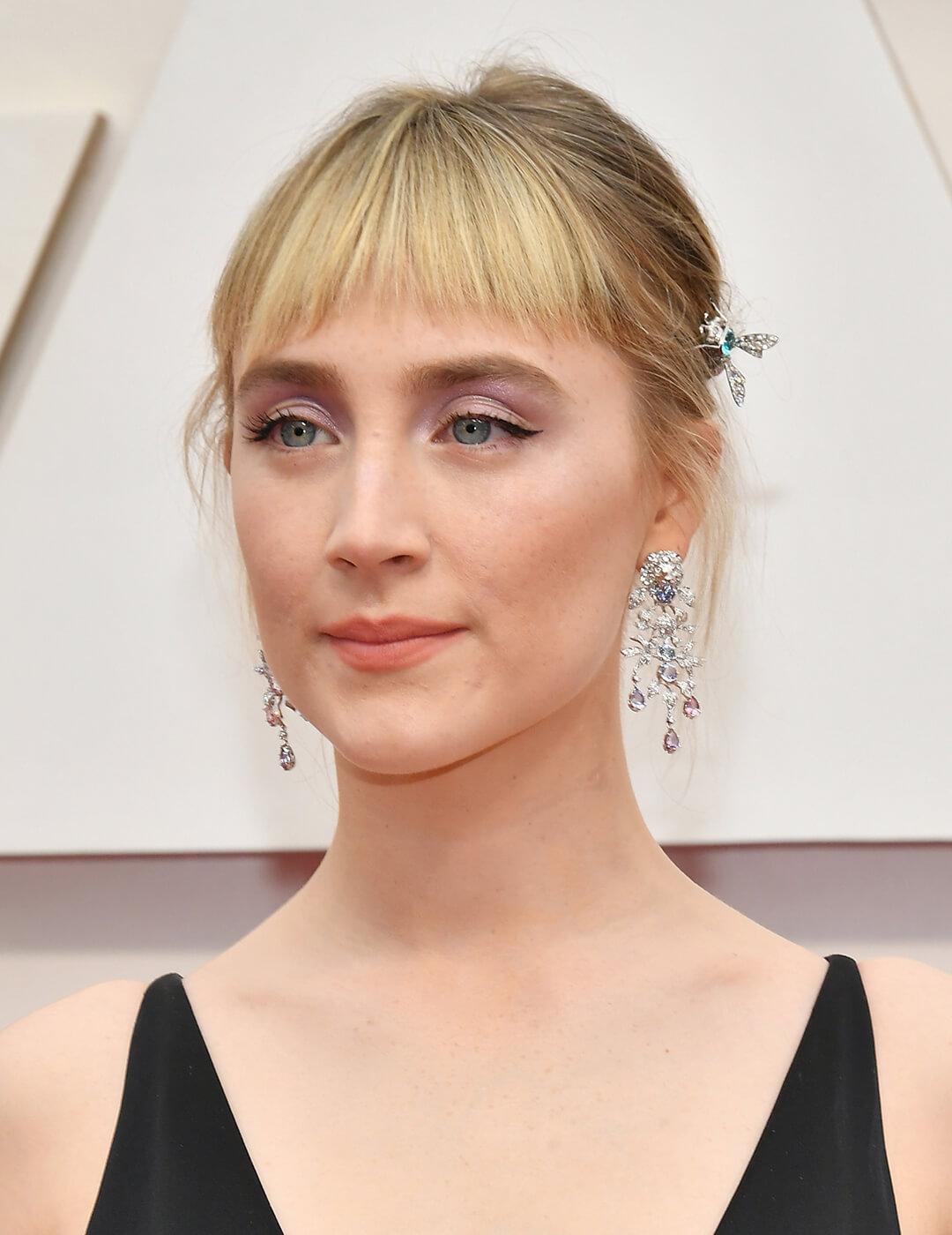 Saoirse Ronan looking elegant in silver jewelry, black dress, lavender eyeshadow makeup look, and updo hairstyle with bangs