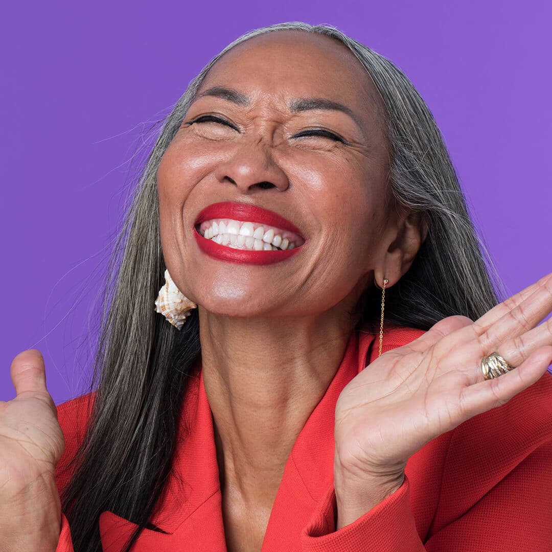 A photo of a mature woman laughing wearing red lipstick and sporting a red coat and conch earrings on a purple background