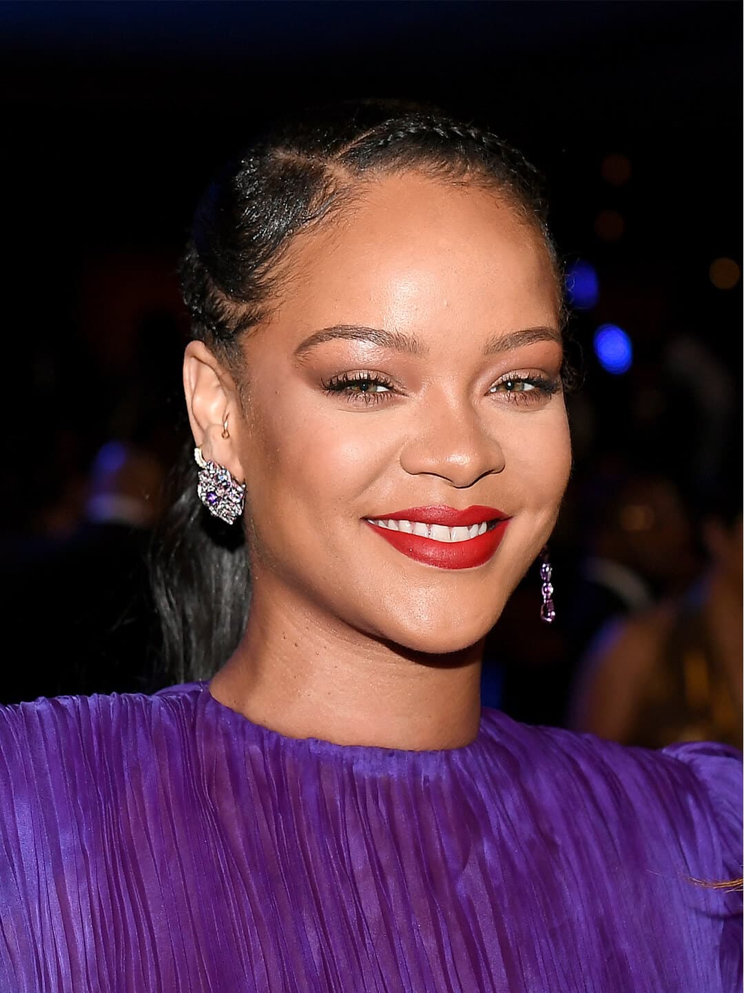 Smiling Rihanna in a purple pleated dress rocking a braided pony tail hairstyle