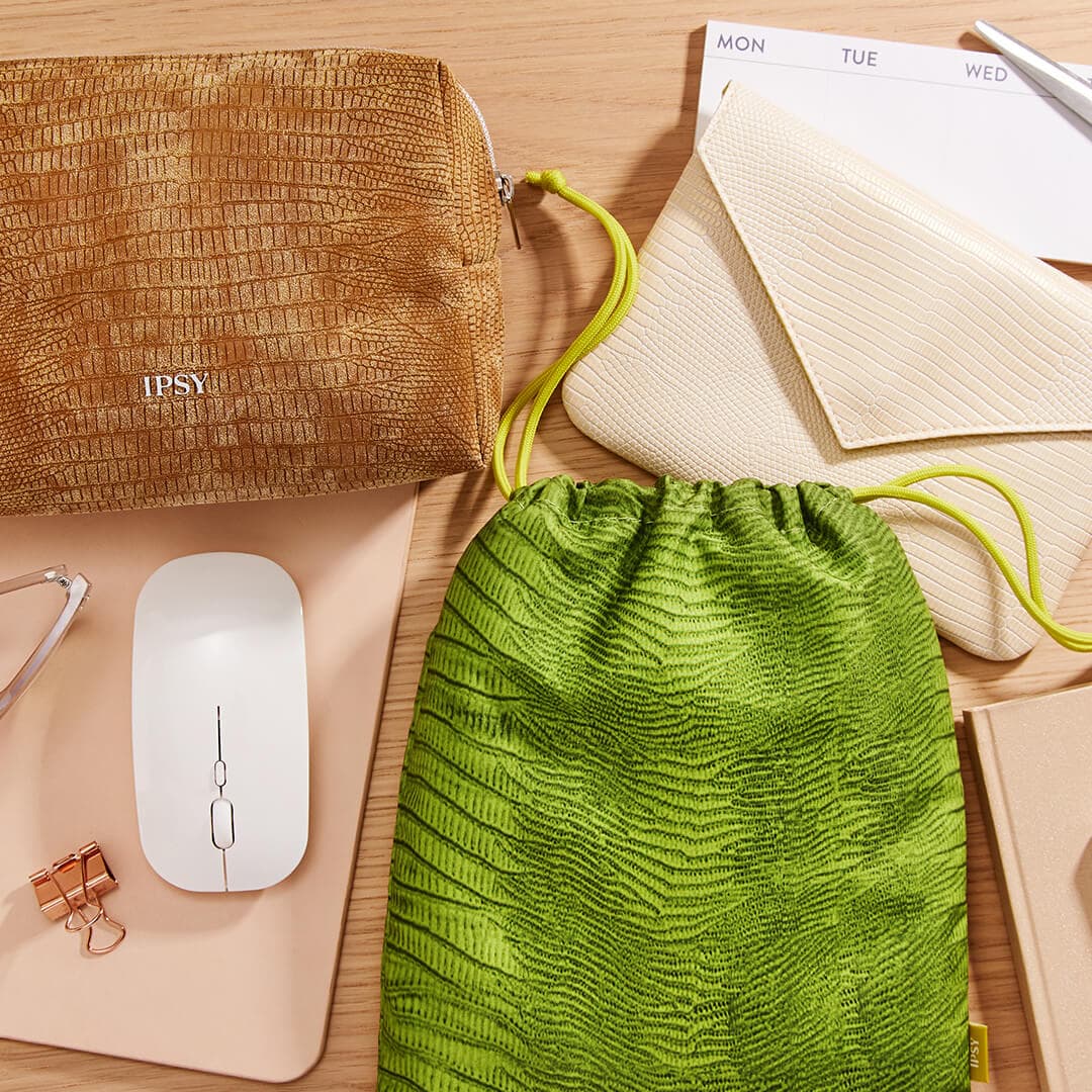 Flat-lay image of the August 2021 IPSY Glam Bag, Glam Bag Plus, and Glam Bag X bags together with a wireless mouse, paper clip, notebooks, and calendar