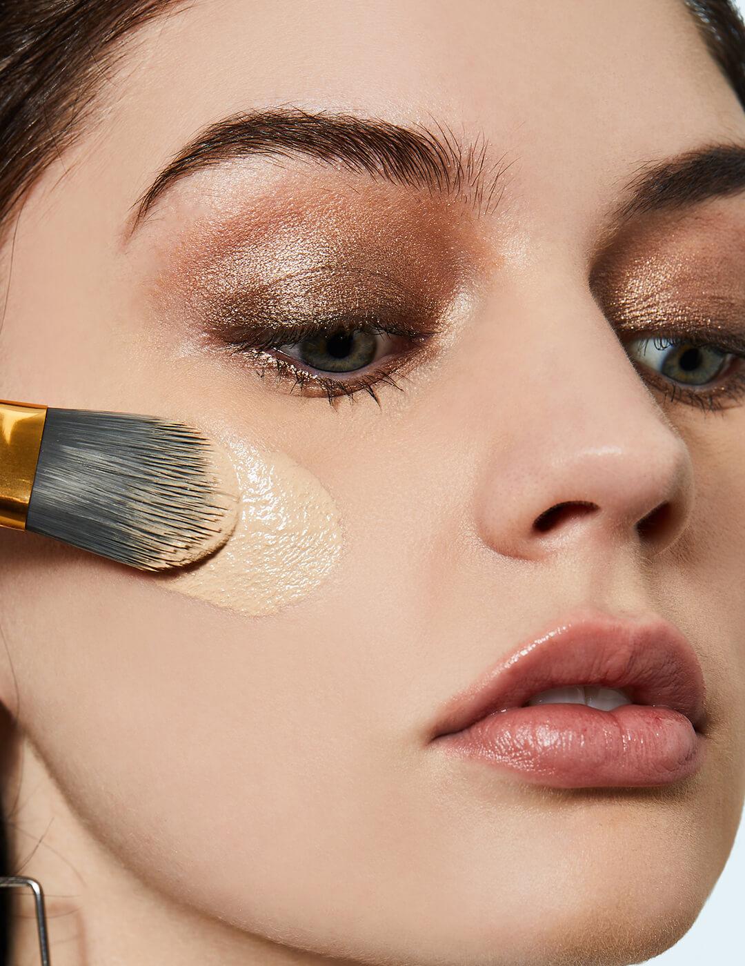 Close-up of a model applying applying foundation on her cheeks while wearing a shimmery nude eyeshadow