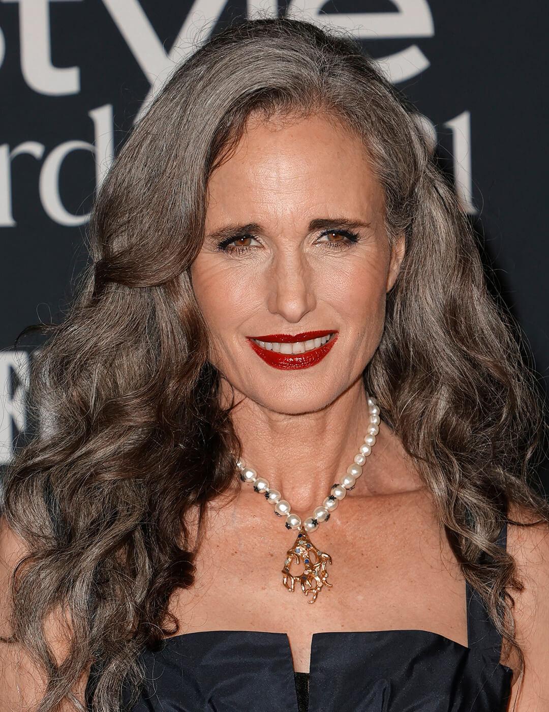 A photo of Andie MacDowell with a smokey eye makeup and red lip look wearing a black dress and a pearl necklace