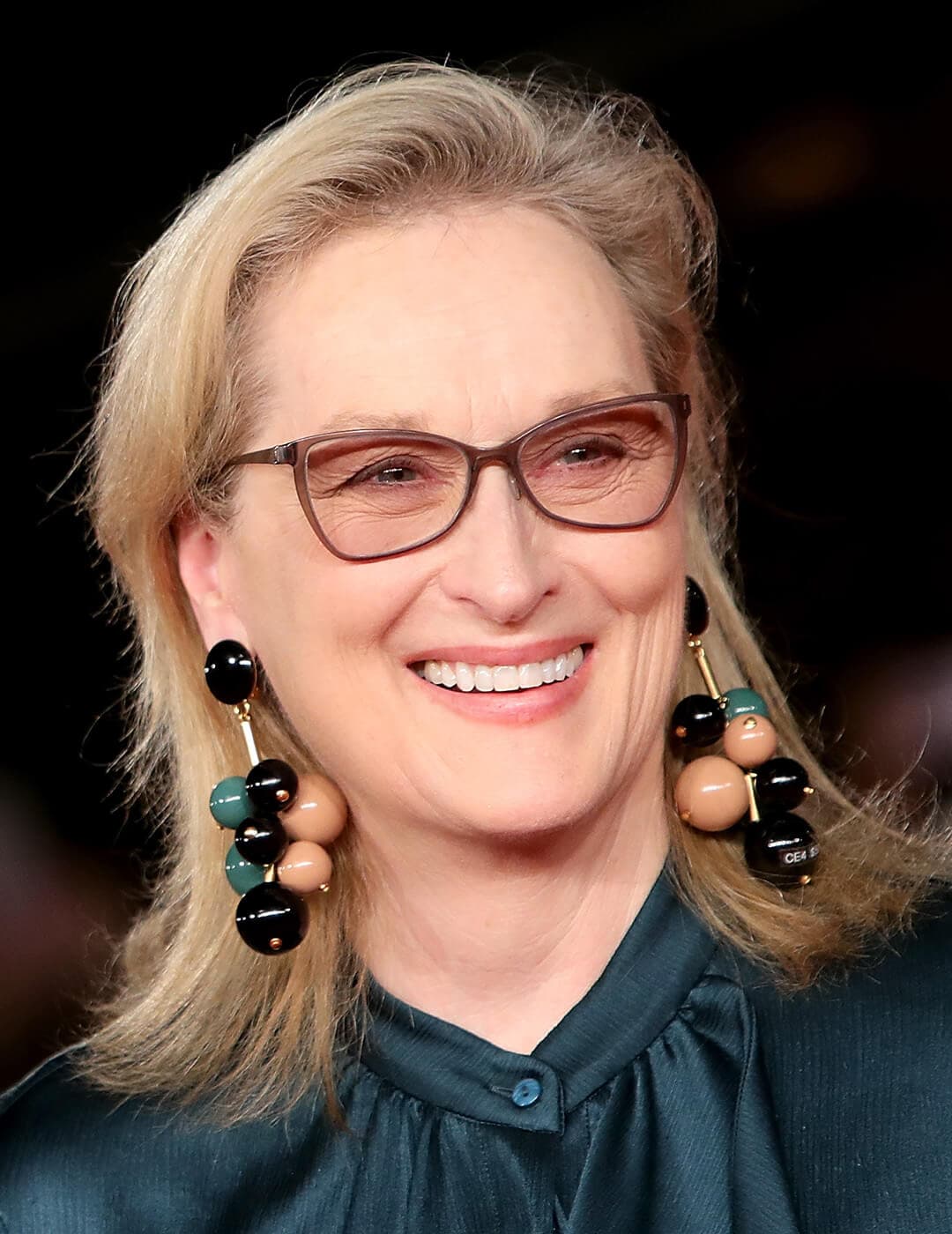 A photo of Meryl Streep wearing brown glasses, ball-drop earrings and a dark green blouse