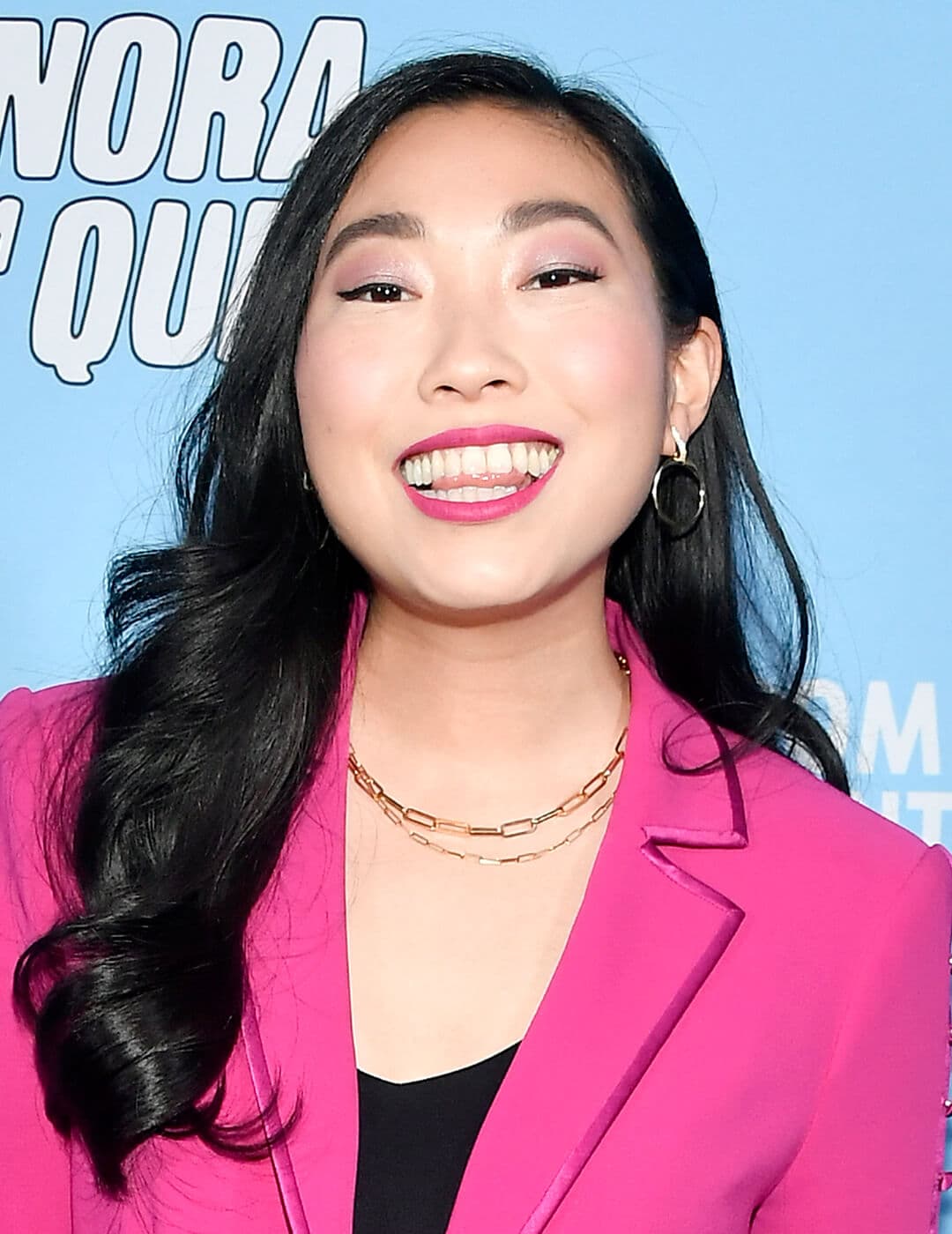 Cheerful Awkwafina rocking a pink suit, black v-nect top, gold necklaces, and wavy hairstyle on the red carpet