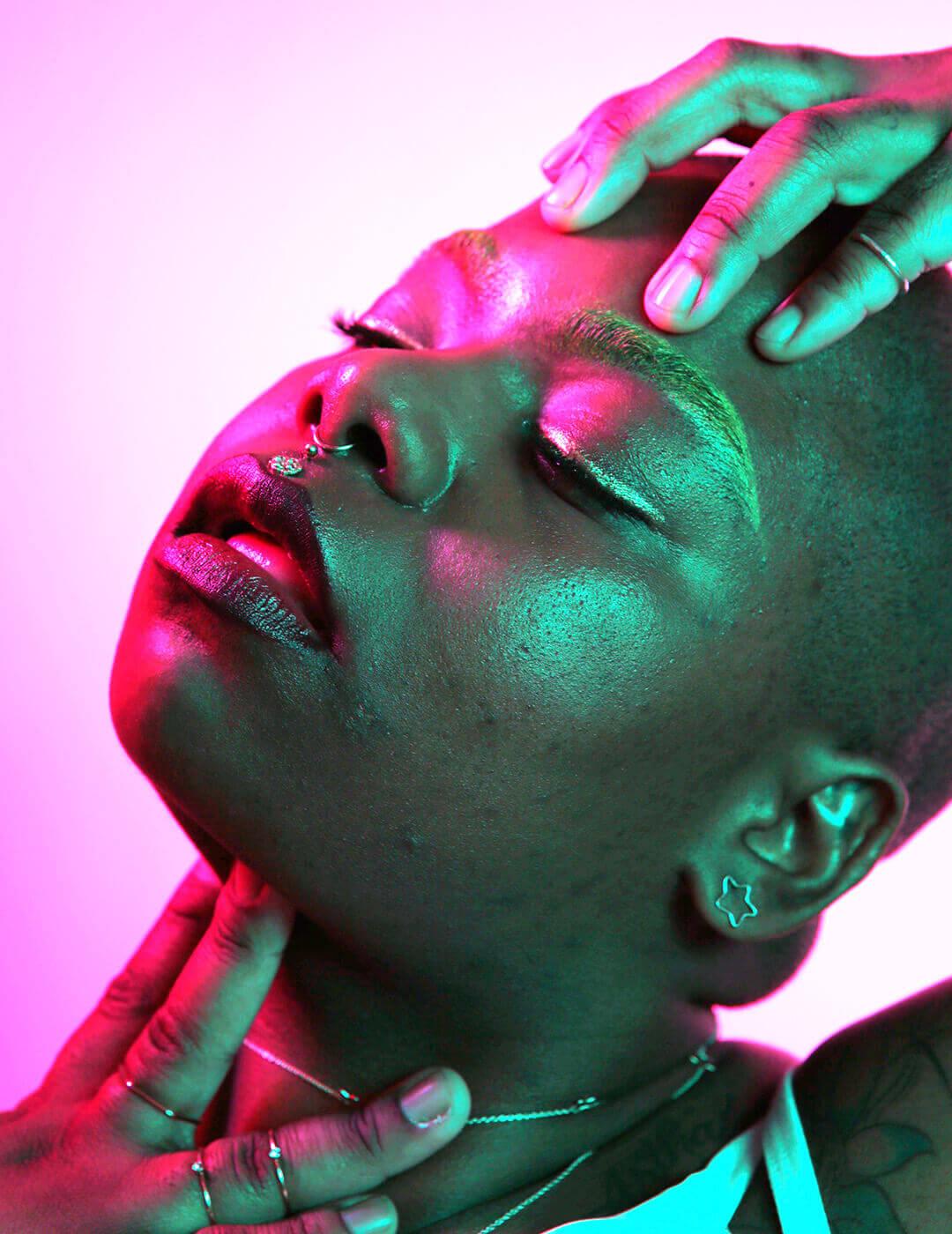 A close-up photo of a woman with a shaved head, her head slightly tilted upward, set against a background illuminated in a soft pink hue infused with AI-driven skincare