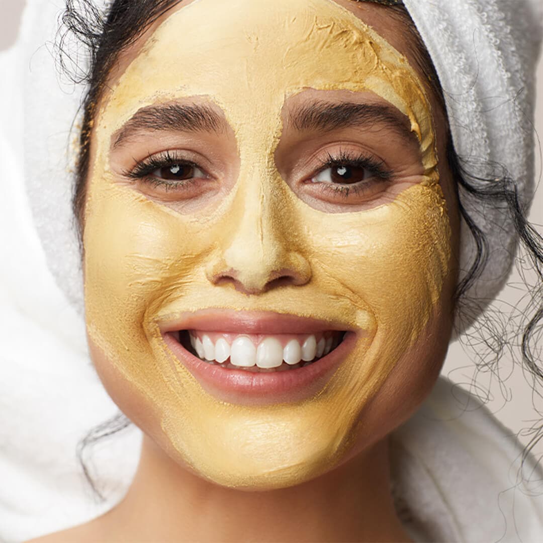 Close-up of a smiling model with her hair wrapped in a towel and yellow face mask on her face