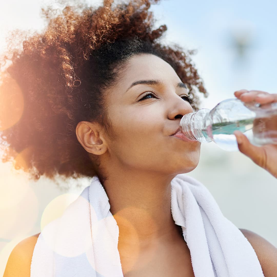 A photo of a woman with a white towel on her neck drinking from a bottle of water