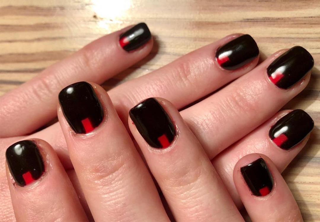A closeup photo of a model's hand with a mix of red on black nail polish on a wooden background