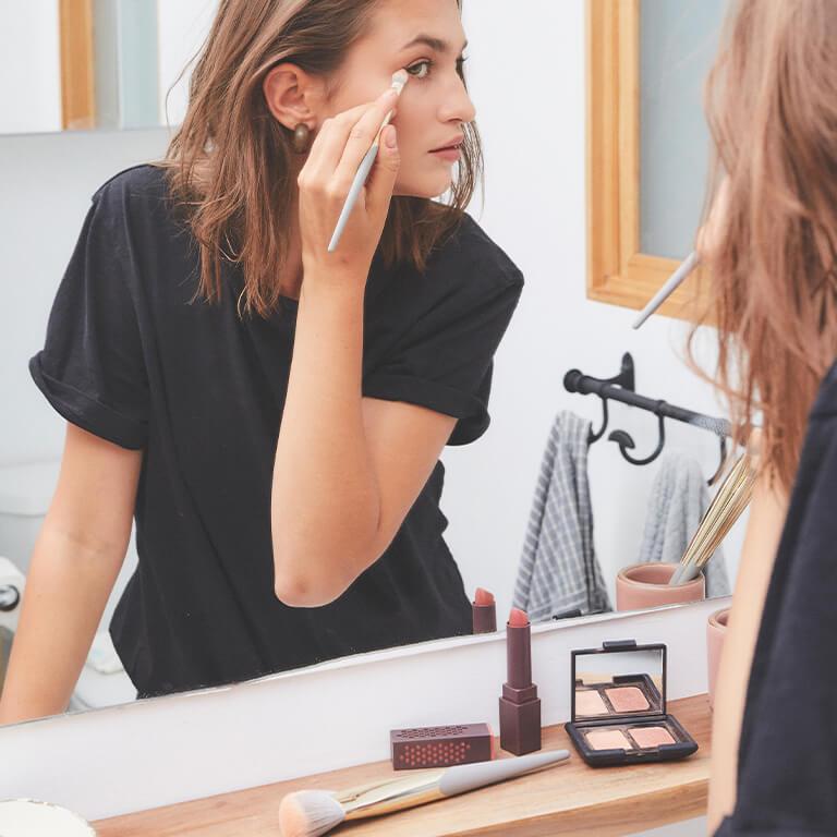 An image of a model in black shirt looking at herself in the mirror while using a COMPLEX CULTURE brush to apply eye makeup